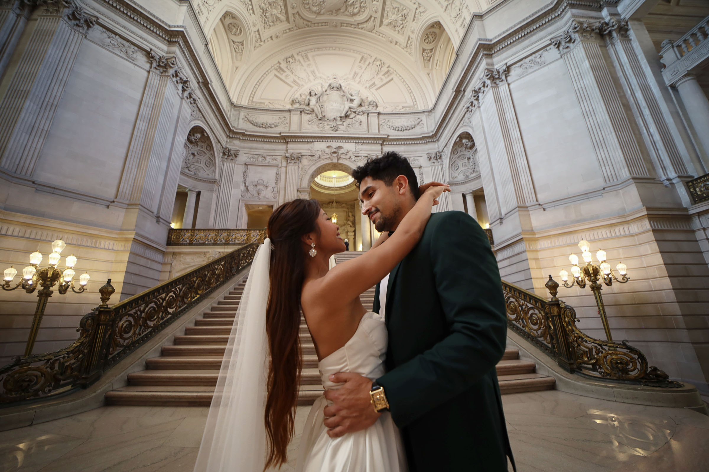 Trinique in a white gown and veil embracing Juan Pablo in a dark green suit at the foot of the grand staircase in San Francisco City Hall